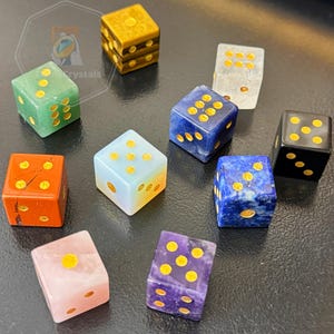Pode incluir: Uma coleção de dados de pedras preciosas em várias cores, incluindo verde, laranja, azul e roxo. Cada dado apresenta pontos dourados. Os dados estão dispostos sobre uma superfície escura, mostrando suas texturas e tons únicos.