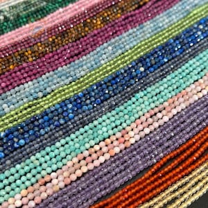 Pode incluir: Uma foto em close-up de vários fios de contas de pedras preciosas em várias cores. As contas são pequenas e facetadas, criando um efeito brilhante. As cores incluem tons de rosa, azul, verde, roxo e laranja. As contas são enfiadas, prontas para fazer joias.