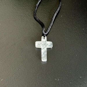 Peut inclure: Un pendentif clair en forme de croix est suspendu &agrave; un collier en cordon noir. Le pendentif a une surface textur&eacute;e et est suspendu sur un fond sombre. Le collier est un bijou simple et &eacute;l&eacute;gant.