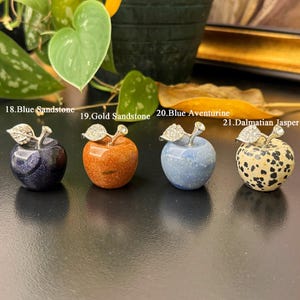 May include: Four decorative apple figurines made of different stones: blue sandstone, gold sandstone, blue aventurine, and Dalmatian jasper. Each apple has a silver-colored stem and leaf design. The apples are displayed on a dark surface.