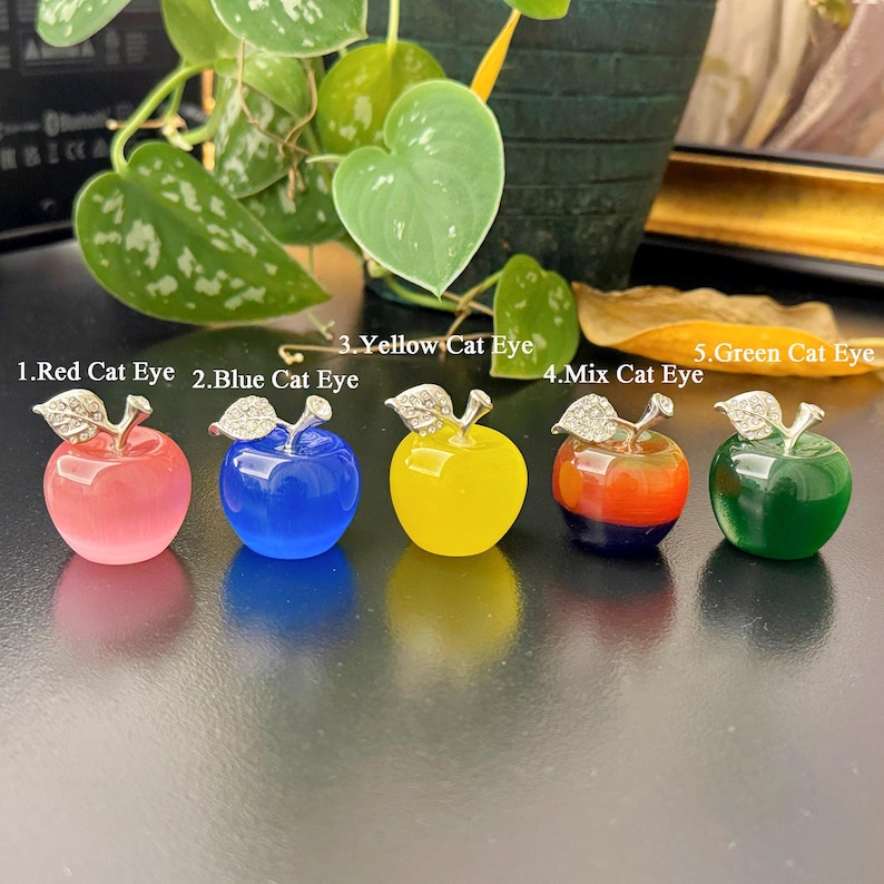 May include: Five colorful apple-shaped ornaments. The ornaments are in shades of red, blue, yellow, orange, and green. Each apple has a silver-colored stem and leaf design. The text on the image reads: "1. Red Cat Eye, 2. Blue Cat Eye, 3. Yellow Cat Eye, 4. Mix Cat Eye, 5. Green Cat Eye."