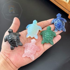 May include: Five small carved stone turtle figurines in various colours including black, translucent white, pink, green, and blue. Each turtle features a detailed shell pattern with a head, four flippers, and a tail, resting on a flat palm surface.