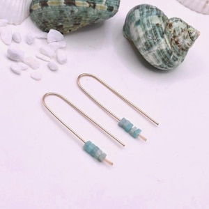 May include: Gold-coloured drop earrings with a linear design, featuring small, light blue gemstone beads. The earrings are displayed with seashells and white pebbles on a white surface, creating a coastal theme.