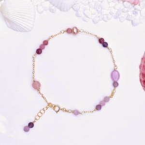 Puede incluir: Delicada pulsera de cadena dorada adornada con una variedad de piedras preciosas rosas y moradas, incluyendo cuentas redondas y ovaladas. Una sola perla está centrada en la pulsera. La pulsera se exhibe sobre una superficie blanca con conchas marinas.