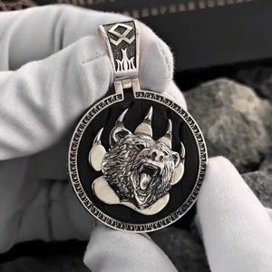 Puede incluir: Colgante de plata con un diseño detallado de cabeza de oso. El oso está gruñendo, rodeado de sus garras, sobre un fondo negro. El colgante se sujeta con una fianza de plata con un diseño intrincado.