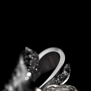 May include: A silver ring with a sculpted design featuring a woman's face and sparkling accents. The ring has a curved, open design and is set against a black background, highlighting its intricate details.