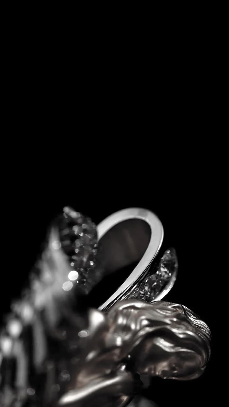 May include: A silver ring with a sculpted design featuring a woman's face and sparkling accents. The ring has a curved, open design and is set against a black background, highlighting its intricate details.