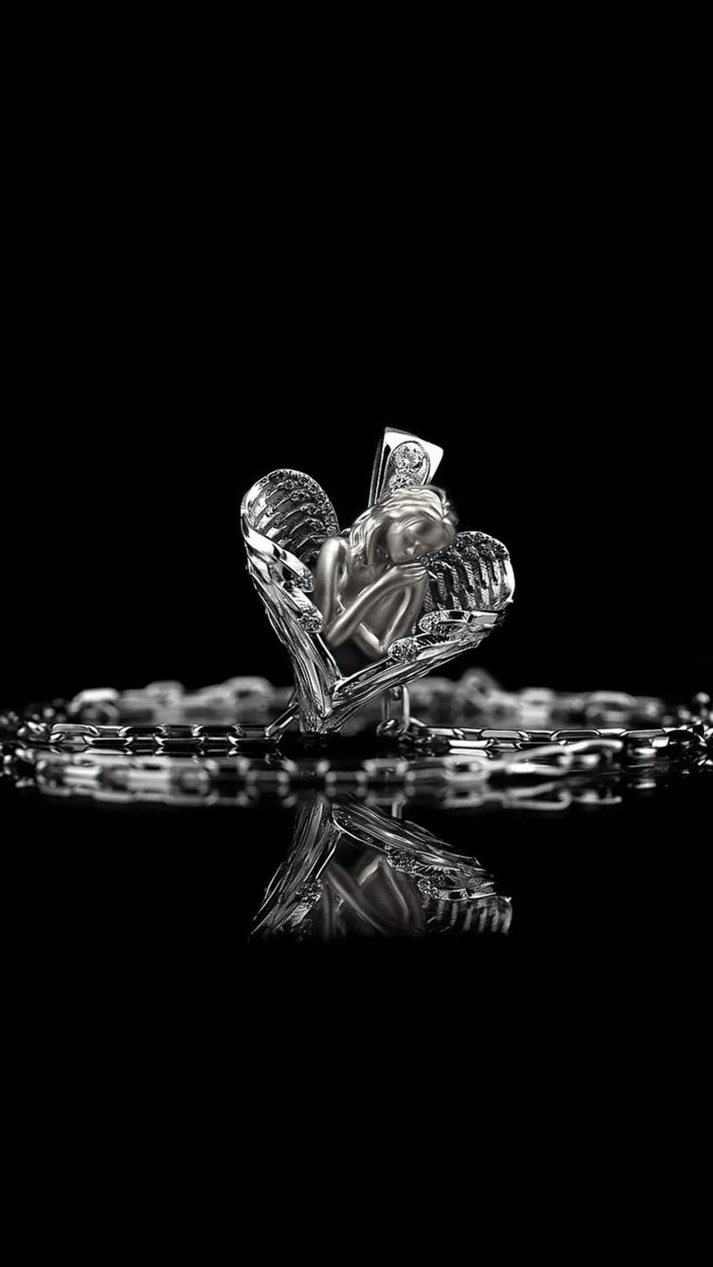 May include: A silver pendant necklace with a detailed angel figure within a heart-shaped frame. The angel has closed eyes and feathered wings. The necklace chain is visible below the pendant, set against a black background.