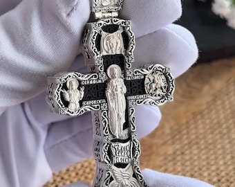 May include: Ornate silver cross with religious figures and inscriptions. The cross features detailed engravings of saints and angels, with text in a stylized font. The design includes a central figure and decorative borders.