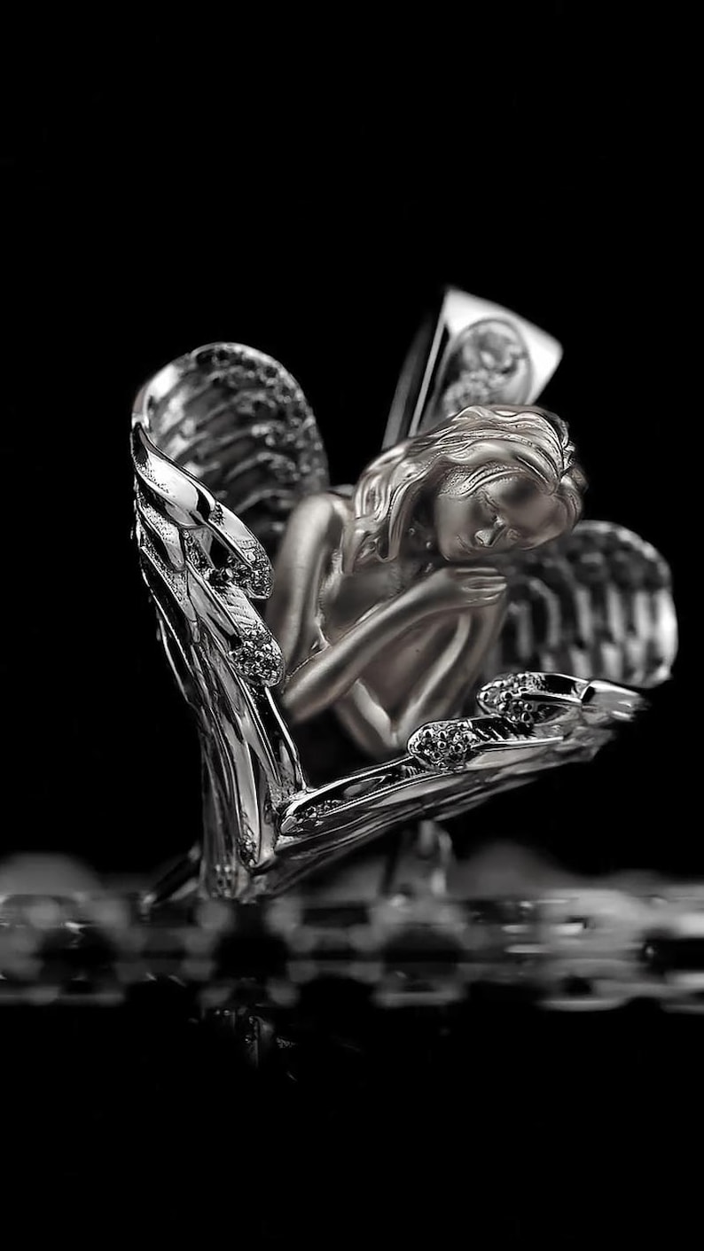 May include: A silver sculpture of an angel, with detailed wings and a serene expression. The angel is cradled within a heart-shaped structure, adorned with sparkling accents. The artwork is set against a dark background.