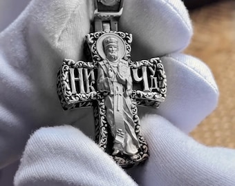 Puede incluir: Colgante de plata con forma de cruz. El colgante presenta una detallada representación de una figura religiosa, con texto cirílico en los brazos de la cruz. El colgante está sujeto por una mano enguantada.