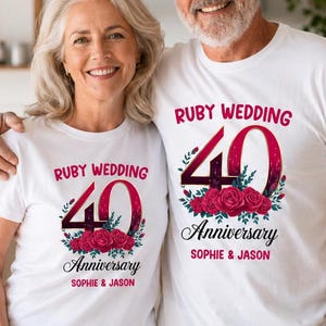 Op de afbeelding: Twee witte t-shirts met een grafisch ontwerp met de tekst "RUBY WEDDING 40 Anniversary" in rood en zwart, vergezeld van een tros rode rozen en groene bladeren, met de namen "SOPHIE & JASON" in het rood onderaan gedrukt.