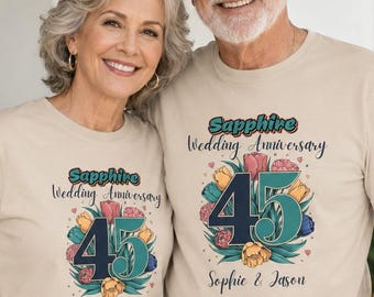 May include: Two beige t-shirts featuring a graphic print with the text Sapphire Wedding Anniversary, the number 45, and a floral arrangement. The design includes the names Sophie & Jason printed below the flowers in a dark script font.