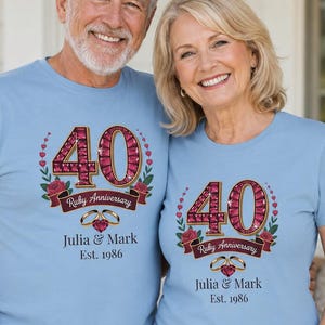 Pode incluir: Duas t-shirts azul-claras com um design gráfico do número 40 em estilo rubi, rodeado por folhas verdes e rosas vermelhas. Abaixo, uma faixa diz Ruby Anniversary, com dois anéis dourados e o texto Julia & Mark Est. 1986.