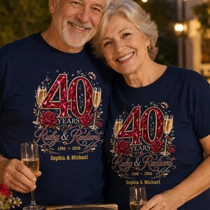 Op de afbeelding: Twee marineblauwe t-shirts met een afbeelding met de tekst 40 YEARS, Ruby & Radiant, 1986-2026 en Sophia & Michael. Het ontwerp bevat een groot rood robijn-getextureerd getal 40, twee champagneglazen en rode rozen. Een bijpassend bordje is aanwezig.