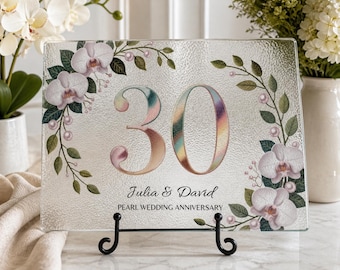 May include: A rectangular glass plaque featuring the number 30 in a metallic iridescent finish, surrounded by printed white orchid flowers, green leaves, and small pearl accents. The text reads Julia & David and PEARL WEDDING ANNIVERSARY.