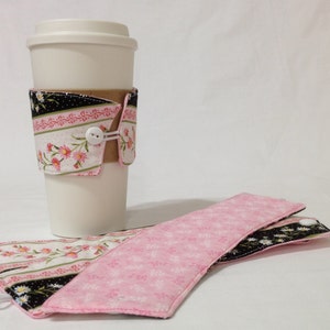 May include: Two pink and black fabric coffee cup sleeves with a white button closure. One sleeve is on a white coffee cup.