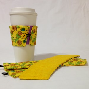 May include: A set of three yellow fabric coffee cup sleeves. One sleeve has a purple and yellow floral pattern with a black button. The sleeves are folded and laid out on a white surface.