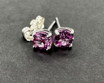 May include: A pair of stud earrings featuring round, faceted, purple gemstones set in silver-colored metal. The earrings are displayed on a dark gray surface, showcasing the sparkling gems and the intricate details of the earring backs.