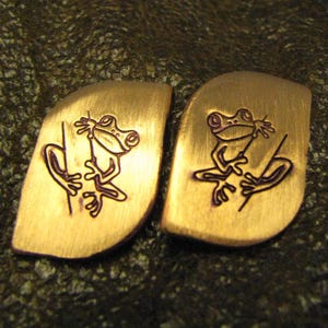 May include: Two copper-colored metal pendants with a frog design. Each pendant has a stylized frog illustration etched in black, clinging to a branch. The pendants have a curved, irregular shape.