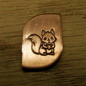 May include: A metal stamp with a cute squirrel design. The squirrel is holding an acorn and is etched into a curved, rectangular piece of metal. The metal has a brushed finish and a warm, copper tone.