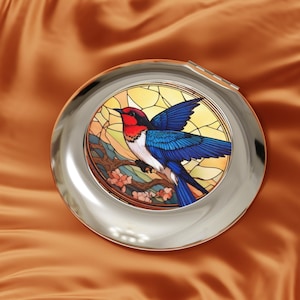 以下が含まれることがあります： カラフルな鳥のデザインが施された、丸い銀色のコンパクトミラー。鳥は赤、青、白の羽を持ち、ピンクの花が咲く枝にとまっています。背景は、黄色と茶色の色合いのステンドグラス風です。