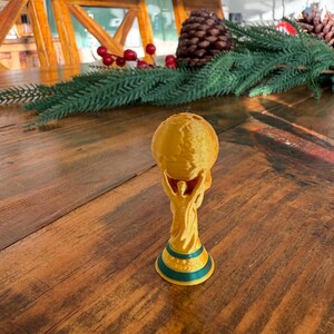 Pu&ograve; includere: Un trofeo dorato con un globo in cima, sorretto da due figure. La base ha una striscia verde e blu. Il trofeo &egrave; su un tavolo di legno. Decorazioni natalizie sullo sfondo.
