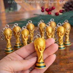 Puede incluir: Llaveros dorados con forma de trofeo de la Copa del Mundo. Los llaveros están impresos en 3D y presentan un diseño detallado del trofeo. La base del trofeo tiene una franja verde y negra. El texto "World Cup Trophy Keychain" es visible.