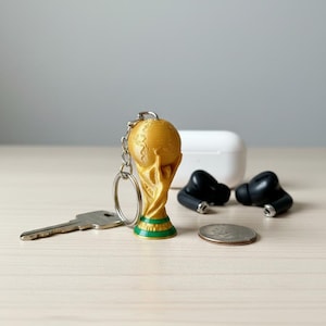 Pu&ograve; includere: Un portachiavi a forma di trofeo dorato con base verde, una chiave d'argento, una moneta e auricolari wireless neri. Sullo sfondo, una custodia di ricarica bianca.