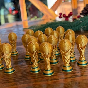 Pu&ograve; includere: Una collezione di statuette trofeo color oro con basi verdi e oro, disposte su una superficie di legno. I trofei presentano una parte superiore a forma di globo e un corpo curvo.