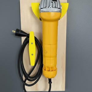 May include: A yellow and silver angle grinder mounted on a wooden board with a yellow holder for the black power cord. The grinder is designed for cutting and grinding materials. The cord is approximately 60 inches long.