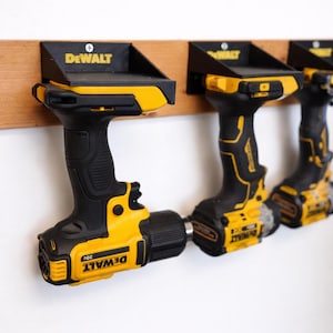 May include: Yellow and black cordless drills mounted on a wooden board. The drills are labelled "DEWALT" and "20V". The drill holders are black and mounted on a light brown wooden board. The background is a plain white wall.