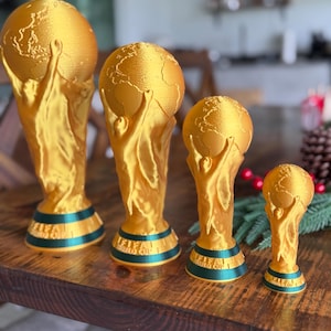 Puede incluir: Cuatro trofeos dorados de la Copa Mundial de la FIFA de diferentes tamaños se exhiben sobre una superficie de madera. Cada trofeo presenta un globo terráqueo sostenido por dos figuras, con una banda verde y azul en la base. Los trofeos están dispuestos de mayor a menor.