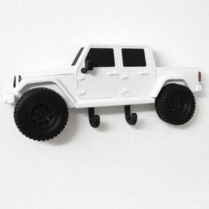 Könnte beinhalten: Weißer, an der Wand montierter Hakenhalter in Jeep-Form mit schwarzen Reifen und Fenstern. Der Halter hat zwei schwarze Haken zum Aufhängen von Gegenständen. Ein dekorativer und funktionaler Artikel.