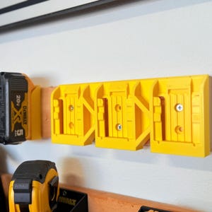 May include: A yellow plastic power tool battery holder mounted on a wall. The holder is designed to hold multiple batteries and is attached to a wooden board. Several black and yellow power tool batteries are visible.