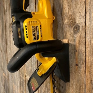 May include: A yellow and black DeWalt 20V MAX cordless hedge trimmer mounted on a wooden wall. The trimmer features a black handle, a yellow body with the DeWalt logo, and a blade guard.