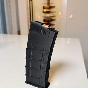 May include: A black magazine with the letters "AAA" printed on the top. The magazine is loaded with several bullets. The magazine is rectangular with a series of rectangular indentations on the side.