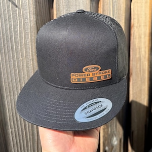 May include: Black snapback hat with a mesh back and a flat brim. The hat features the Ford logo and the words "POWER STROKE DIESEL" in orange. The hat is held by a person.