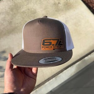 May include: A brown and white trucker hat featuring a rectangular orange patch on the front panel with the black text 6.7L POWER STROKE TURBO DIESEL. The hat has a flat brim and a circular sticker on the brim that reads THE AUTHENTIC SNAPBACK.