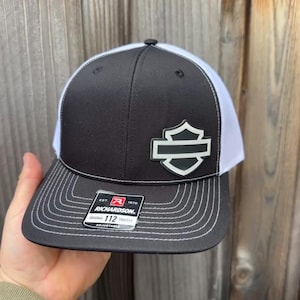 May include: Black and white trucker hat with a shield logo. The hat features a black front panel, a white mesh back, and a curved brim with white stitching. The Richardson brand tag is visible.
