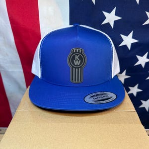 May include: A blue and white trucker hat with the Kenworth logo on the front. The hat has a blue brim and crown, a white mesh back, and a snapback closure. The hat is sitting on a cardboard box.
