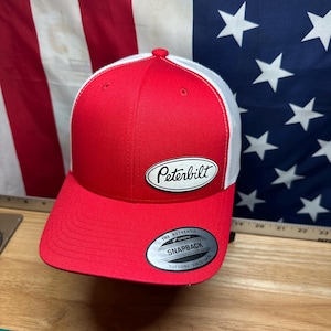 May include: A red and white trucker hat with the Peterbilt logo on the front. The hat has a curved brim and a white mesh back. The hat has a snapback closure and the text "The Authentic Snapback Yupoong Since 1974".