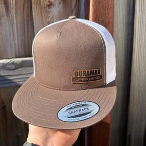 May include: Brown and white trucker hat with a brown Duramax Turbo Diesel logo patch. The hat has a brown brim and a white mesh back. The hat also has a white sticker on the bottom of the brim that says "The Authentic Yupoong Snapback Yupoong Since 1974."