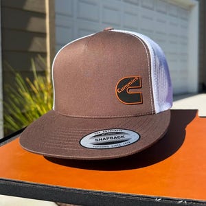 May include: Brown and white trucker hat with the Cummins logo on the front. The hat has a brown brim and front panel, a white mesh back, and a snapback closure. The Cummins logo is in orange and black.