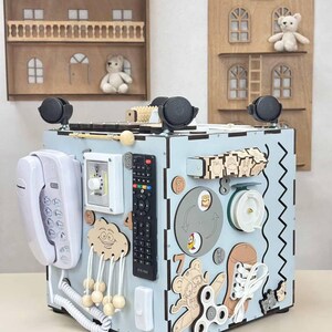 May include: A light blue wooden activity cube for toddlers, featuring various interactive elements. Includes a toy phone, remote control, spinning toys, and wooden shapes. The cube has black wheels and is decorated with cartoon animal figures.
