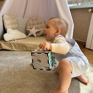 Könnte beinhalten: Ein Kind spielt mit einem Holz-Aktivitätswürfel. Der Würfel hat ein Zifferblatt, einen Riegel und andere interaktive Elemente. Der Würfel ist weiß mit schwarzen und türkisfarbenen Akzenten. Das Kind trägt eine hellblaue Latzhose.