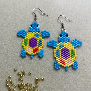 May include: Handmade beaded turtle earrings. The earrings feature a blue head and flippers, a yellow shell with a purple center, and accents of red, green, and pink. The earrings are on a gray surface with gold beads.