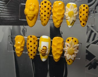May include: A set of ten yellow press-on nails with various designs. Some nails feature a textured surface, while others have black polka dots. Two nails are adorned with a 3D bee and a flower design. The nails are displayed on a clear plastic holder.