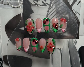 May include: A set of ten artificial nails with various designs. Some nails feature a clear base with a pink French tip, while others showcase red flowers, green leaves, and ladybug accents. The nails are arranged on a clear plastic display.