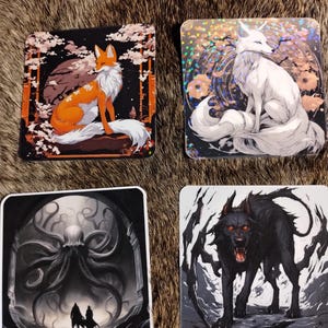 Peut inclure: Quatre dessous de verre carrés avec de l'art fantastique. L'un représente un renard orange avec une queue blanche, un autre un renard blanc avec une longue queue, un troisième une scène en noir et blanc avec des personnages et le dernier un loup noir aux yeux rouges.